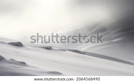 Traces of wind on the surface of the snow-covered slope of the mountain. The slope of the mountain in the background is hidden in the winter haze. Royalty-Free Stock Photo #2064568490