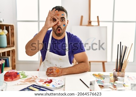 Young hispanic man with beard at art studio with painted face doing ok gesture shocked with surprised face, eye looking through fingers. unbelieving expression. 