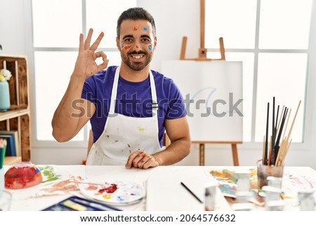 Young hispanic man with beard at art studio with painted face smiling positive doing ok sign with hand and fingers. successful expression. 