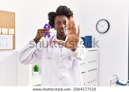African doctor woman holding purple ribbon awareness at medical clinic with open hand doing stop sign with serious and confident expression, defense gesture 