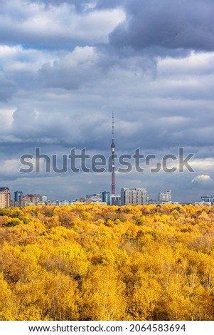 dark gray rainy clouds in blue sky over lush yellow forest illuminated by sun and a city with TV tower on horizon on autumn day before rain