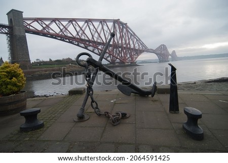 A picture of a sea anchor monument in front of a red rail bridge crossing a mysterious lagoon