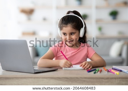 Distance Education. Little Arab Female Child Wearing Wireless Headphones Study With Laptop At Home, Smiling Cute Girl Drawing With Pencils In Notepad And Looking At Computer Screen, Free Space