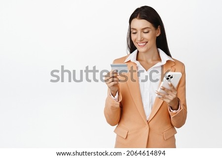Smiling young businesswoman paying online, holding credit card and smartphone, order on phone, standing over white background