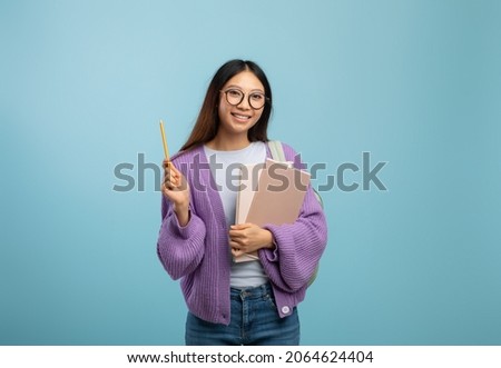 I know the answer. Excited asian female student in eyeglasses holding notepad and pointing pencil up, wearing backpack while standing over blue studio background