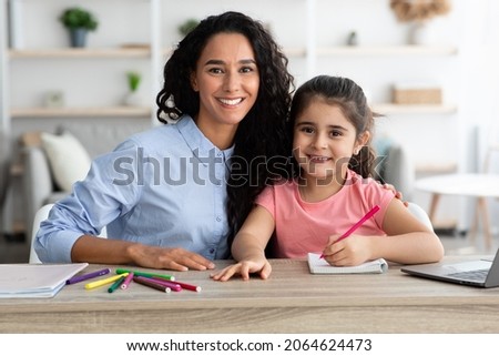 Portrait Of Happy Young Mother And Little Daughter Posing At Table While Making School Homework Together At Home, Caring Arab Mom Helping Her Cute Female Child With Study, Smiling At Camera