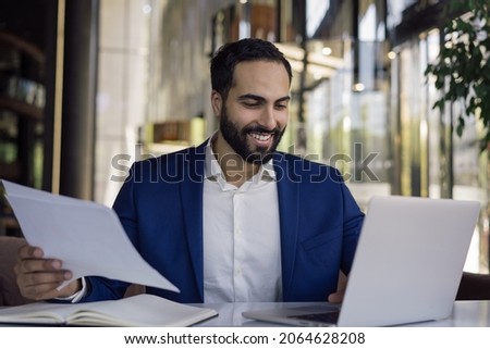 Handsome smiling middle eastern businessman using laptop computer  planning startup project. Successful arab manager working with business documents, holding financial report sitting in modern office