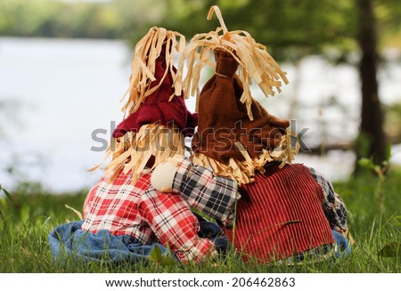 Boy and girl scarecrows back view sitting in grass by lake