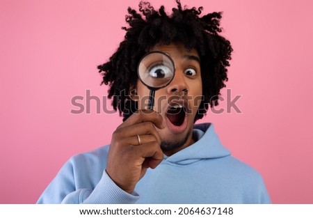 Shocked black teen guy looking through magnifying glass on pink studio background. Surprised African American teenager suspecting, investigating or questioning something. Secret, doubt concept Royalty-Free Stock Photo #2064637148