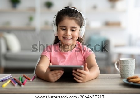 Cute Little Arab Girl Wearing Wireless Headphones Relaxing With Digital Tablet At Home, Adorable Female Child Watching Cartoons On Tab Computer While Sitting At Table In Living Room, Closeup