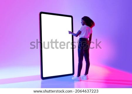 Millennial African American woman touching screen of huge tablet computer, interacting with pc interface in neon light, mockup for new app or website design. Touch pad template Royalty-Free Stock Photo #2064637223