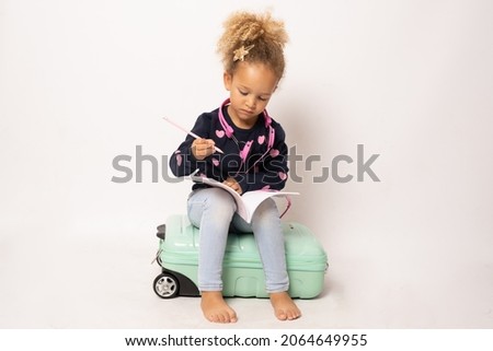 Cute little girl sits on a suitcase writing in a notebook isolated over white background.