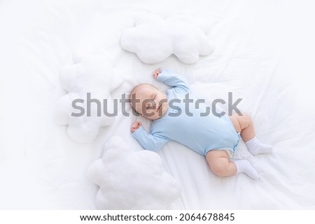 baby boy sleeps on the bed lying on his back in blue pajamas with his hands up among the pillows of clouds, healthy newborn sleep Royalty-Free Stock Photo #2064678845