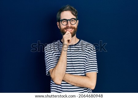 Caucasian man with beard wearing striped t shirt and glasses looking confident at the camera with smile with crossed arms and hand raised on chin. thinking positive.  Royalty-Free Stock Photo #2064680948