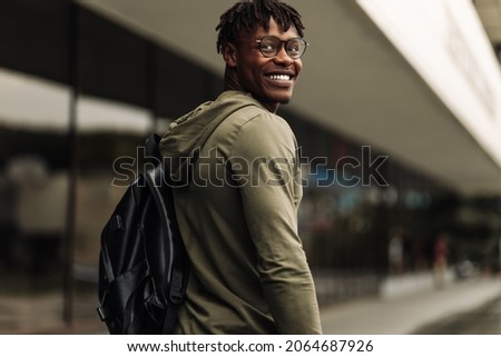 Happy handsome successful african student, wearing glasses and with a black backpack, standing on the steps and smiling, looks into the frame outdoors on the street near the university