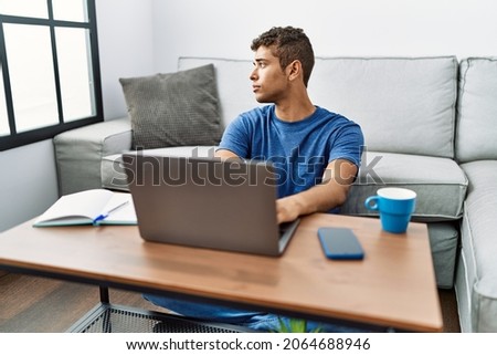 Young handsome hispanic man using laptop sitting on the floor looking to side, relax profile pose with natural face with confident smile. 