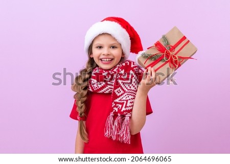 A little girl in a red Christmas scarf and a festive hat holds her Christmas gift and smiles.