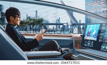 Futuristic Concept: Handsome Stylish Japanese Businessman in Glasses Reading Notebook and Watching News on Augmented Reality Screen while Sitting in a Autonomous Self-Driving Zero-Emissions Car. Royalty-Free Stock Photo #2064717011