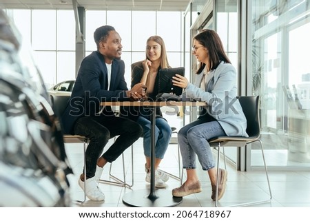 Caucasian saleswoman using digital tablet while working with multi ethnic couple at car salon. Happy man and woman talking with competent manager while buying auto.
