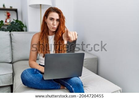 Young redhead woman using laptop at home pointing with finger to the camera and to you, confident gesture looking serious 