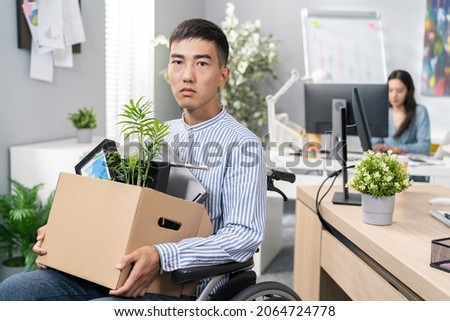 Disabled ambitious man in shirt with Asian Korean beauty sits in wheelchair at desk in the company on knees holding cardboard box with packed things, gets fired from job unable to fulfill his duties Royalty-Free Stock Photo #2064724778