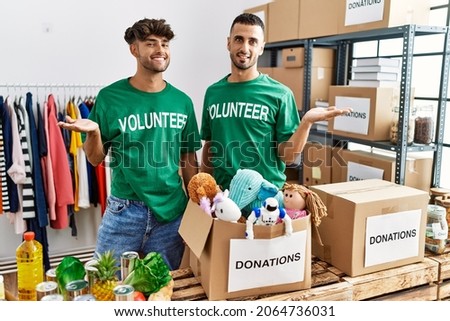 Young gay couple wearing volunteer t shirt at donations stand smiling cheerful presenting and pointing with palm of hand looking at the camera. 