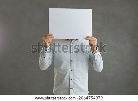 Unknown man on a gray background holding a poster with empty copy space for your message. Person who hides his identity behind a blank white sheet of paper. Concept of anonymity. Banner.