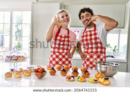 Couple of wife and husband cooking pastries at the kitchen smiling doing phone gesture with hand and fingers like talking on the telephone. communicating concepts. 