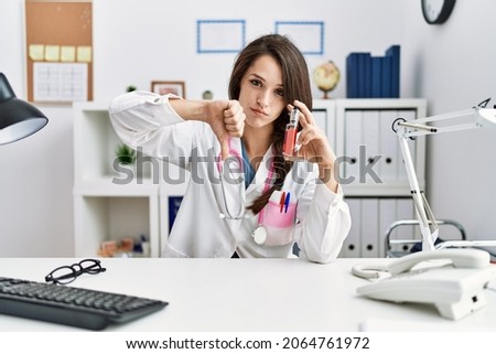 Young doctor woman holding electronic cigarette at medical clinic with angry face, negative sign showing dislike with thumbs down, rejection concept 