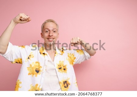 Young blonde man in shirt smiling while making winner gesture isolated over pink background