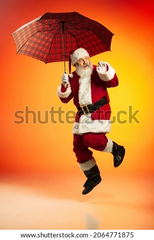 Unpredictable weather. Joyful Santa Claus with umbrella jumping isolated on red yellow background in neon. Happy holiday, wishes, dreams concept, Merry Christmas and Happy New Year 2022
