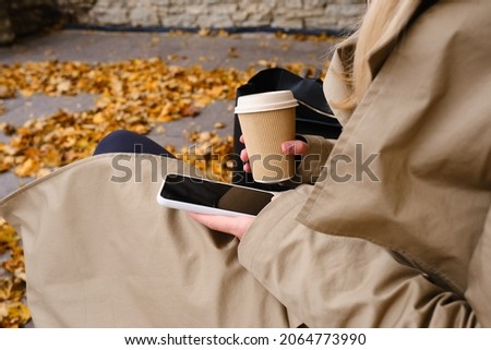 Girl with a smartphone and a cup of coffee sits on a bench in an autumn park. A woman is sending a text message or using an app on her mobile phone. Hot drink in a paper cup for a cozy walk