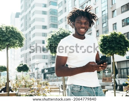 Handsome hipster model.Unshaven African man dressed in white summer t-shirt.Fashion male with dreadlocks hairstyle. Posing in the street.Looking at smartphone screen, using cellphone apps