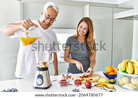 Middle age hispanic couple pouring juice on mixer machine cooking smoothie at the kitchen.
