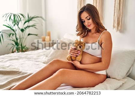 Young pregnant woman in white underwear sits on the bed in the bedroom with a teddy bear. Maternity concept.