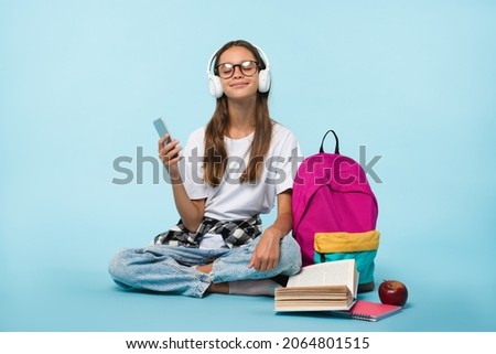 Cheerful caucasian schoolgirl teenager pupil student listening to the music song singer playlist in headphones while preparing going back to school isolated in blue background