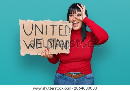 Young hispanic woman holding united we stand banner smiling happy doing ok sign with hand on eye looking through fingers 