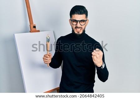 Handsome man with beard holding brushes close to easel stand angry and mad raising fist frustrated and furious while shouting with anger. rage and aggressive concept. 