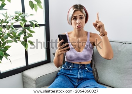 Young beautiful woman using smartphone typing message sitting on the sofa pointing up looking sad and upset, indicating direction with fingers, unhappy and depressed. 