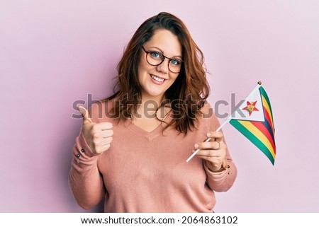 Young caucasian woman holding zimbabwe flag smiling happy and positive, thumb up doing excellent and approval sign 