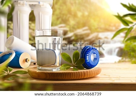 Glass of water purified by osmosis with environmentally friendly biodegradable filters on wooden slat table with nature background trees and waterfall. Front view. Horizontal composition. Royalty-Free Stock Photo #2064868739