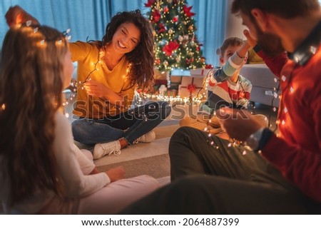 Parents and children having fun spending Christmas Eve together at home, sitting on the floor by the Christmas tree, playing with Christmas lights