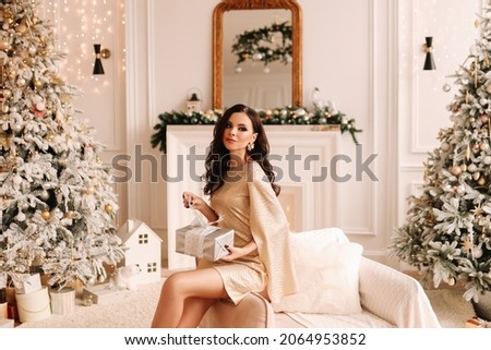 A pretty brunette young woman with a hairstyle in an elegant outfit holds a gift box in the decorated interior of the house during the Christmas holiday and New Year indoor. Selective focus Royalty-Free Stock Photo #2064953852