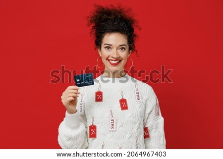 Young smiling happy cheerful rich female costumer woman 20s wear white knitted sweater with tags sale in store showroom hold in hand credit bank card isolated on plain red background studio portrait