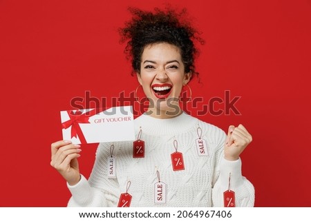 Young happy fun costumer woman 20s wear white knitted sweater with tags sale in showroom hold gift certificate coupon voucher card for store do winner gesture isolated on plain red background studio
