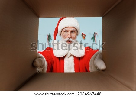 Old impressed bearded Santa Claus man 50s wears Christmas hat red suit clothes look at gifts in paper box isolated on plain blue background studio. Happy New Year 2022 merry ho x-mas holiday concept