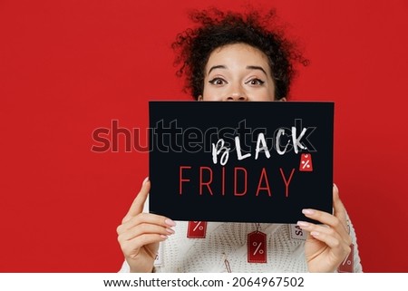 Young caucasian female costumer woman 20s in white knitted sweater with tags sale hold in hand cover mouth with card sign with balck friday title text isolated on plain red background studio portrait