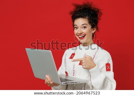 Young smiling fun happy female costumer woman 20s wear white knitted sweater with tags sale in store showroom hold use work point finger on laptop pc computer isolated on plain red background studio
