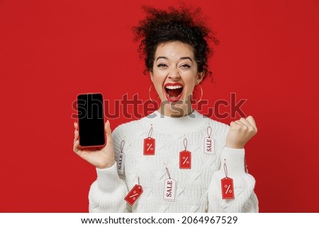 Young woman 20s in white knitted sweater with tags sale in store showroom hold in hand use mobile cell phone with blank screen workspace area do winner gesture isolated on plain red background studio
