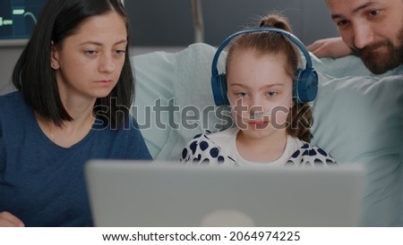 Family sitting beside daughter while playing online videogames using laptop computer in hospital ward during sickness consultation. Sick child recovering after disease infection surgery Royalty-Free Stock Photo #2064974225